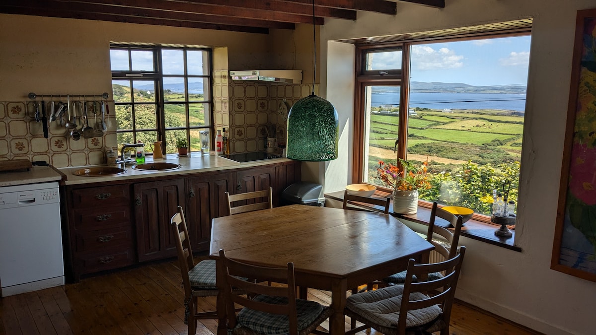 The kitchen features a wooden dining table surrounded by several chairs. A large window provides views of the countryside and sea. Counters are equipped with essential appliances, and decorative elements include a hanging light and plants enhancing the inviting atmosphere.