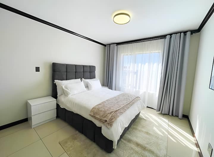Elegant bedroom featuring a neatly made bed with white linen, complemented by a brown accent throw. Natural light filters in through sheer curtains, creating a serene ambiance.