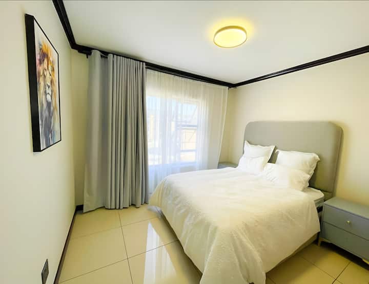 Cozy bedroom with a plush bed dressed in crisp white linen, accented by a light grey headboard. Natural light pours in through sheer curtains, complemented by a warm ceiling light. A painting adds a touch of elegance to the cream-coloured walls.