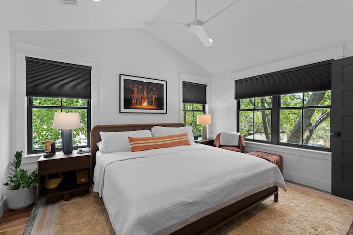 Светла спалня с голямо двойно легло, удобен стол и прозорци с изглед към дървета.