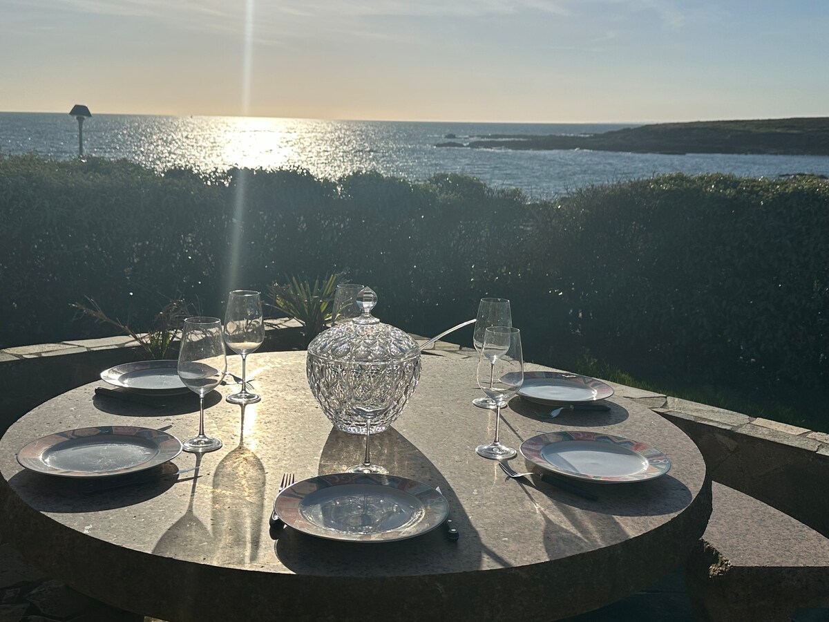 A round table is set for outdoor dining, featuring elegant glassware and fine china placed on a stone surface. The table overlooks a serene view of the ocean, with sunlight reflecting on the water in the background.