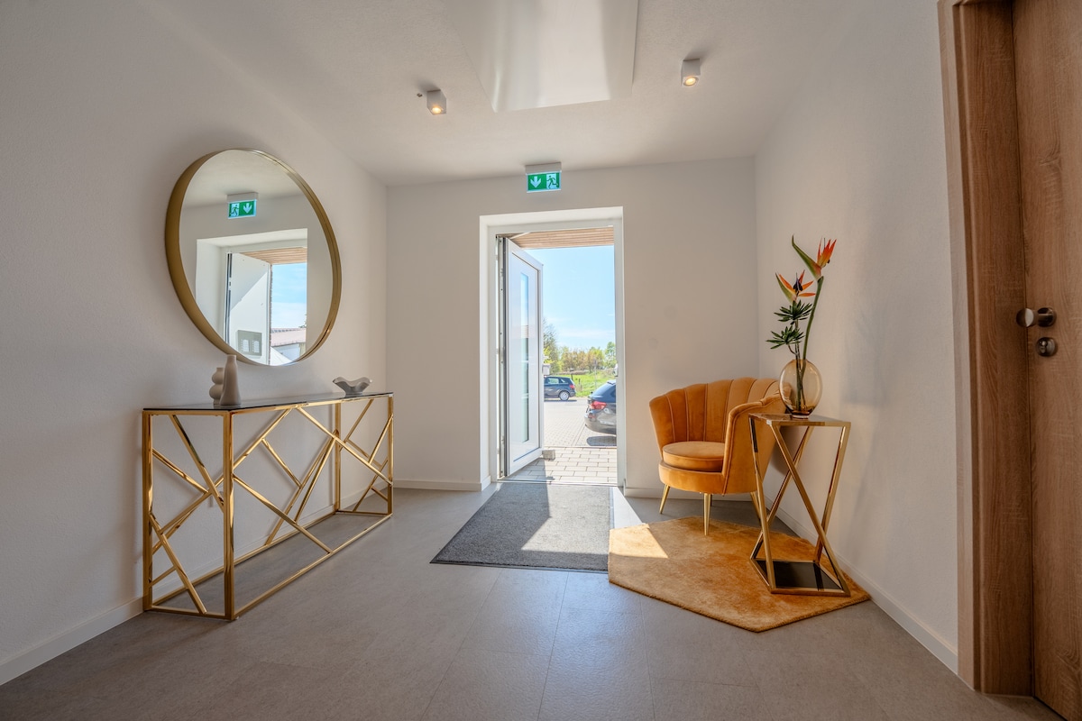 An elegantly designed entryway features a round mirror and a gold geometric console table. A vibrant orange chair is positioned next to a small table adorned with a decorative item. Natural light pours in through the open door, illuminating the spacious area.