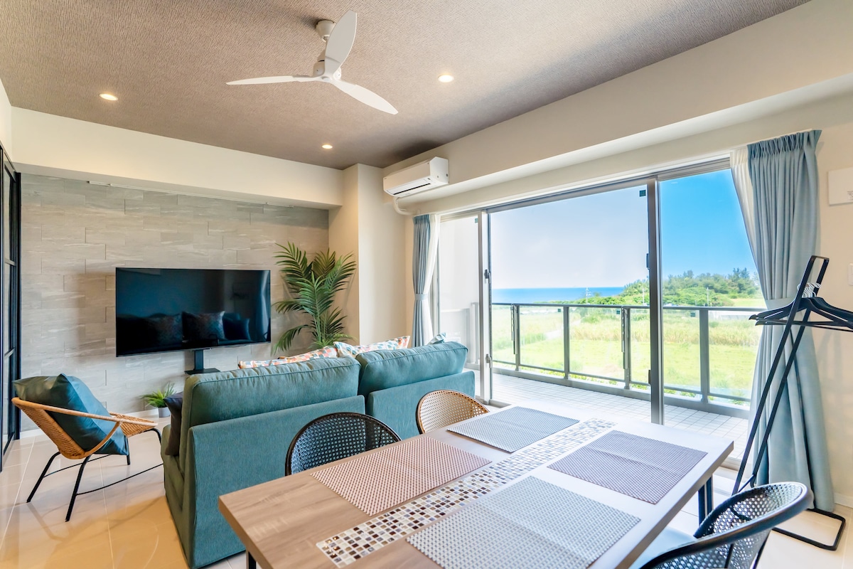 A spacious living area features large glass doors that open to a view of the sea. The room is furnished with a comfortable sofa, a television mounted on the wall, and a dining table surrounded by mesh chairs. Natural light fills the space from the balcony.