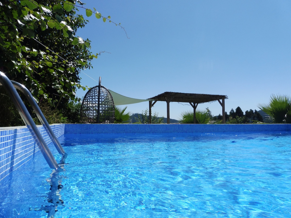 The clear azure pool reflects the bright blue sky, surrounded by leafy greenery. A wooden pergola with a shade sail stands in the background, enhancing the serene outdoor space. Steps lead into the pool, inviting relaxation on warm days.