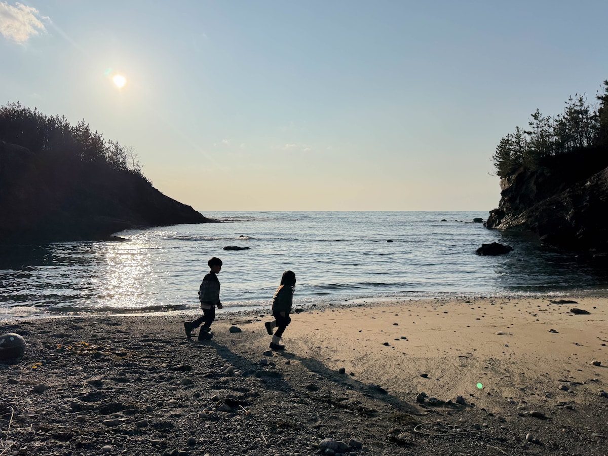 Two children are playing on a sandy beach under soft sunlight. The gentle waves lap at the shore, and distant wooded hills create a serene backdrop. Their silhouettes express joy as they explore the shoreline with a peaceful ocean stretching in the background.