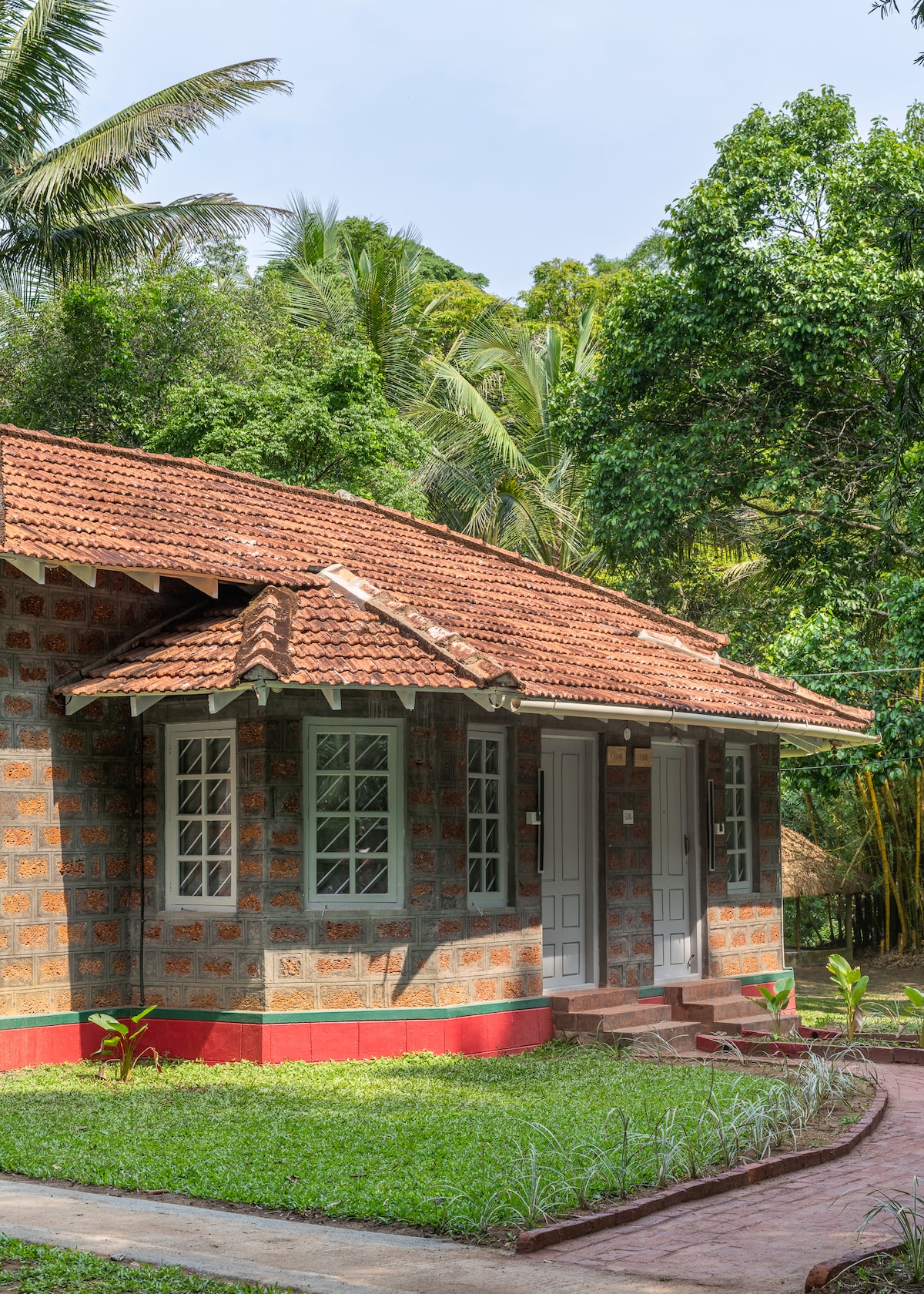 The exterior of Timber Cottage features earthy stone walls and a traditional red-tiled roof, set amidst lush greenery. The two-room structure is surrounded by manicured lawns, with inviting pathways leading to the entrances. Tall trees create a natural backdrop, enhancing the serene atmosphere.