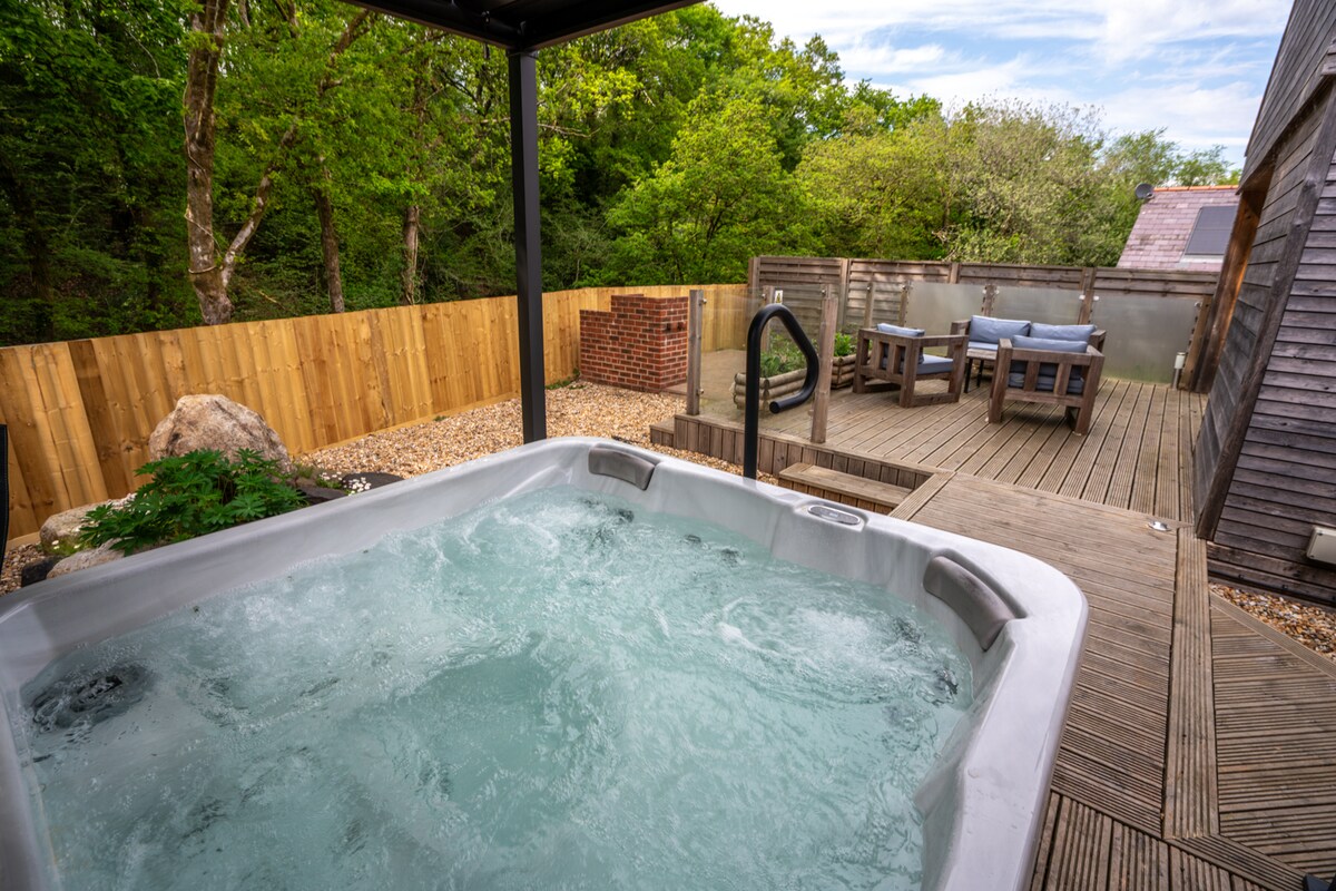 A private hot tub is situated on a wooden deck, surrounded by lush greenery and a wooden fence. Outdoor seating is arranged nearby at a wooden dining table, creating a tranquil space for relaxation.