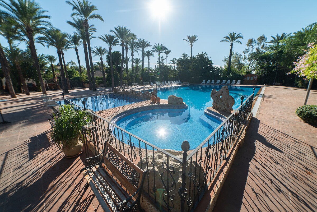 A spacious outdoor pool is surrounded by tall palm trees, creating a relaxed atmosphere. The clear water reflects sunlight, while a stone feature adds a decorative touch. Several lounge chairs are positioned nearby along the tiled patio, inviting visitors to unwind.