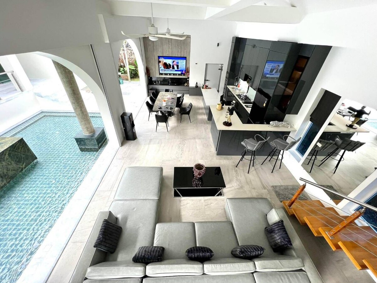 A spacious open-plan living area is visible, showcasing a modern kitchen and dining area. A large Smart TV is mounted on the wall. The inviting lounge features a gray sectional sofa with decorative cushions, alongside a glass coffee table. An outdoor pool is partly visible through large windows.