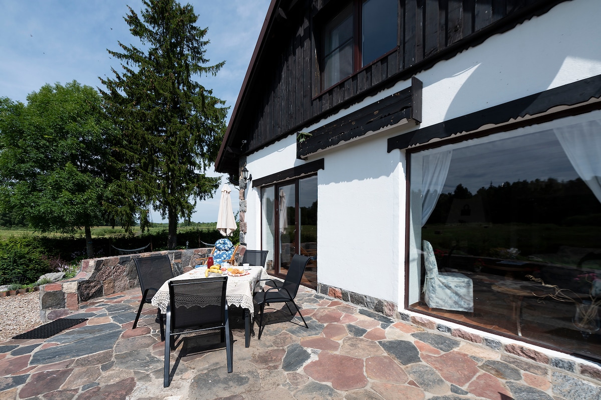 An outdoor stone terrace features a table set with a tablecloth and an assortment of food. Surrounding black chairs and a lounge area can be seen, along with the exterior of the house that includes large windows, allowing for ample natural light.