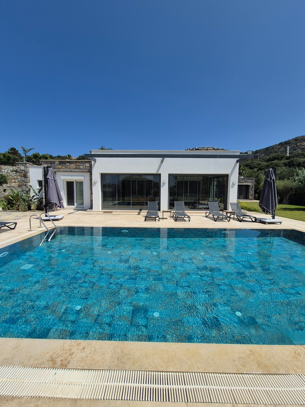 A clear view of a private swimming pool is presented, showcasing vibrant blue water. Several lounge chairs are arranged around the pool area, providing a space for relaxation. The main house is visible in the background, featuring large glass doors that allow for ample natural light.