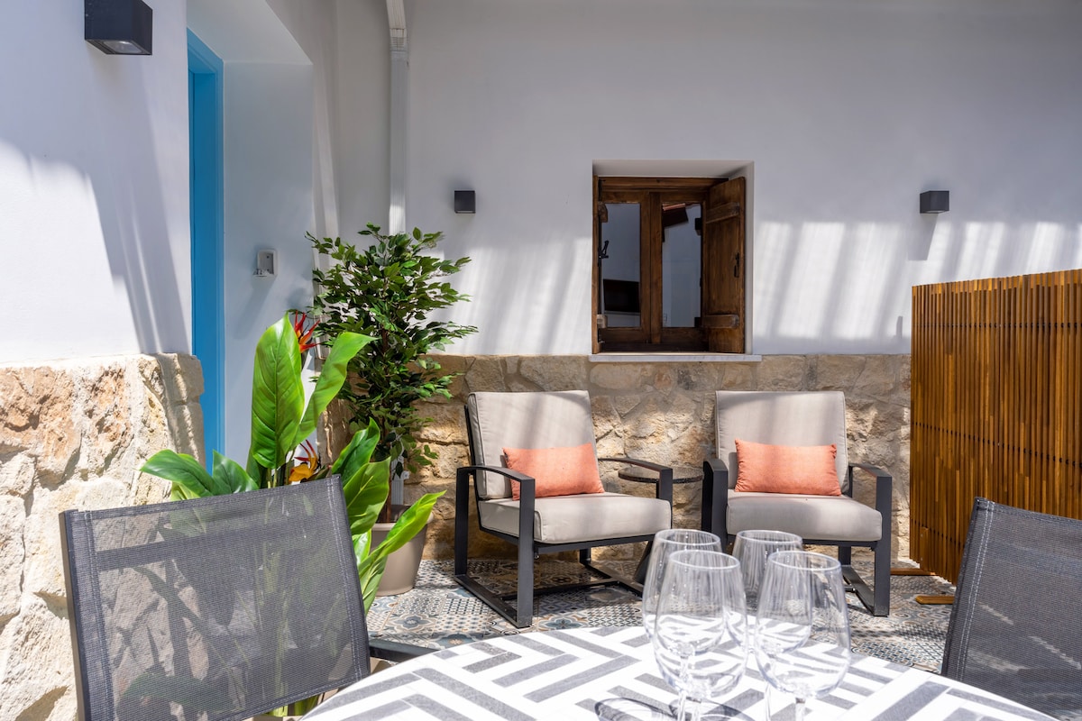 The shared patio features comfortable seating arrangements with two armchairs adorned with orange cushions. A circular dining table is set with glasses, and vibrant green plants add a touch of nature. The stone wall and wooden partition screens provide privacy and a cozy atmosphere.