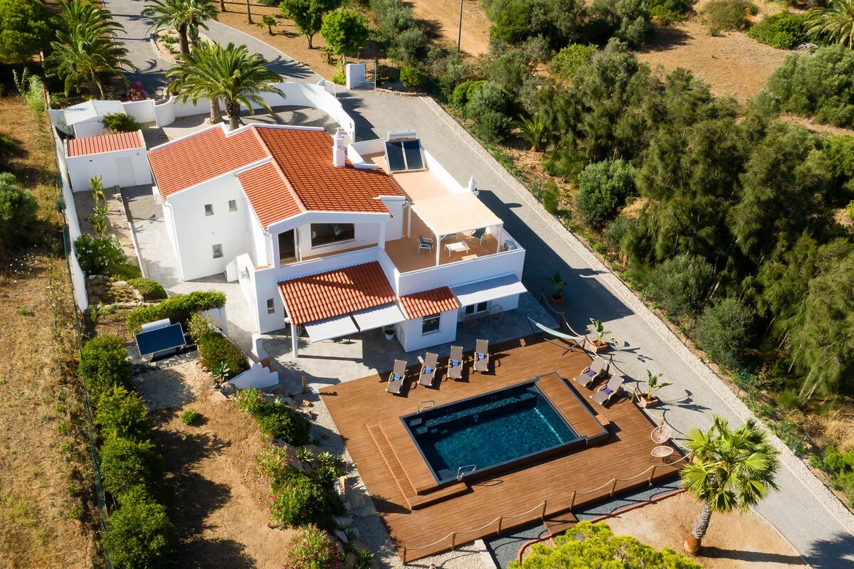 An aerial view of a villa surrounded by greenery shows a spacious layout. A wooden deck features a swimming pool, while palm trees and garden areas enhance the outdoor space. The villa's red-tiled roof adds a charming contrast to the white structure.