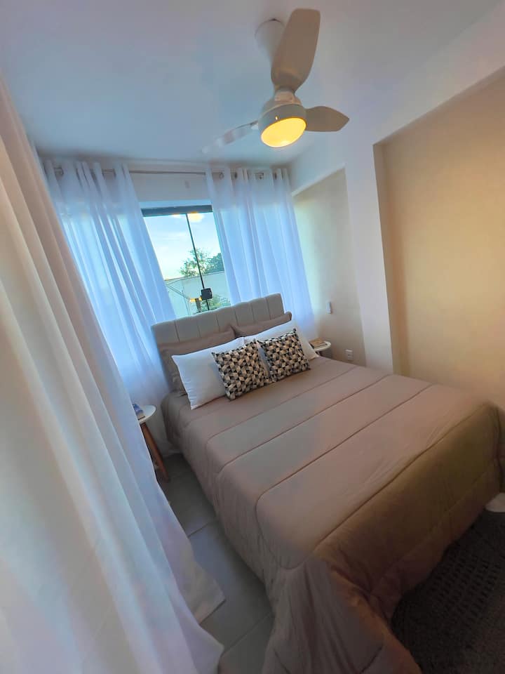 Cozy bedroom with double bed, beige bedding and decorative pillows in neutral tones. Translucent white curtains allow natural light to enter, and a ceiling fan ensures comfort during your stay.