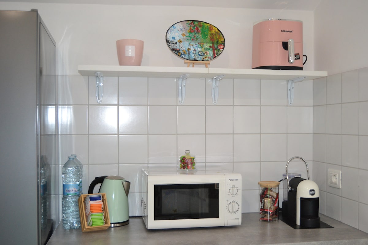 A modern kitchen is displayed with essential appliances such as a microwave, kettle, and coffee machine. Decorative items include a colorful tray and a potted plant, while bottled water is also visible on the countertop, contributing to a functional and inviting cooking space.