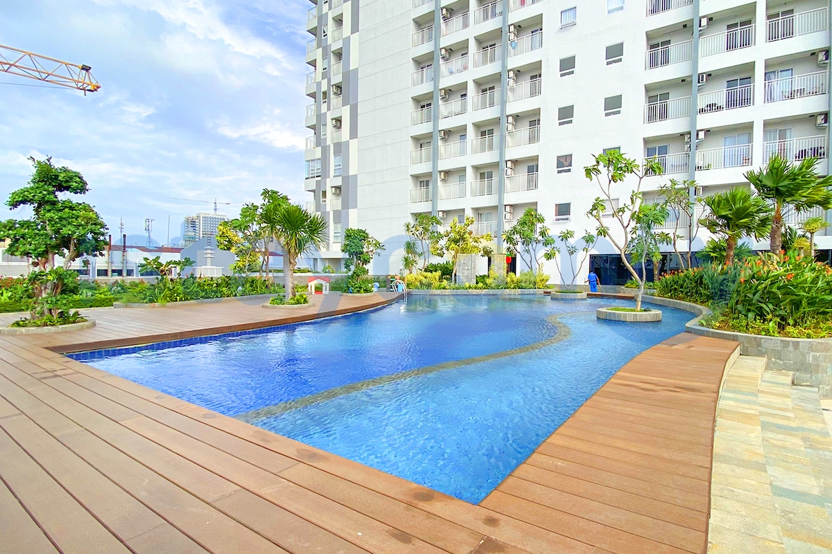 The outdoor pool area is surrounded by landscaped gardens featuring various plants and trees. A wooden deck edges the pool, which reflects the sky. Adjacent buildings appear in the background, enhancing the spacious feel of the area.