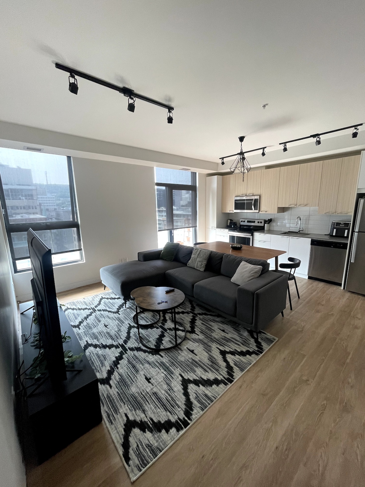 The living area showcases a modern grey sofa arranged around a circular coffee table on a patterned area rug. A large flat-screen TV is positioned against the wall, while the kitchen area is visible behind, featuring sleek cabinetry and essential appliances, all illuminated by natural light from the windows.
