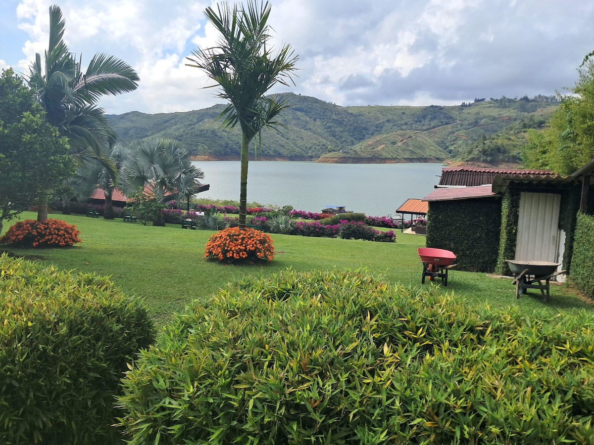 The landscape features a serene lake surrounded by rolling hills, vibrant flowers, and lush greenery. Various structures are nestled among the plants, providing a natural ambiance, while palm trees are positioned throughout the area, enhancing the tranquil outdoor setting.