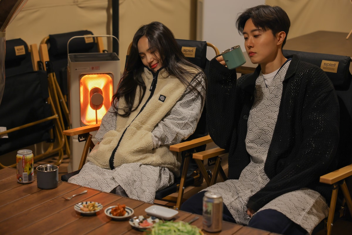 Two individuals are seated on comfortable camping chairs around a wooden table. A small heater emits warmth in the background. An assortment of food items is displayed on the table, including a variety of drinks. The cozy attire enhances the relaxed atmosphere.