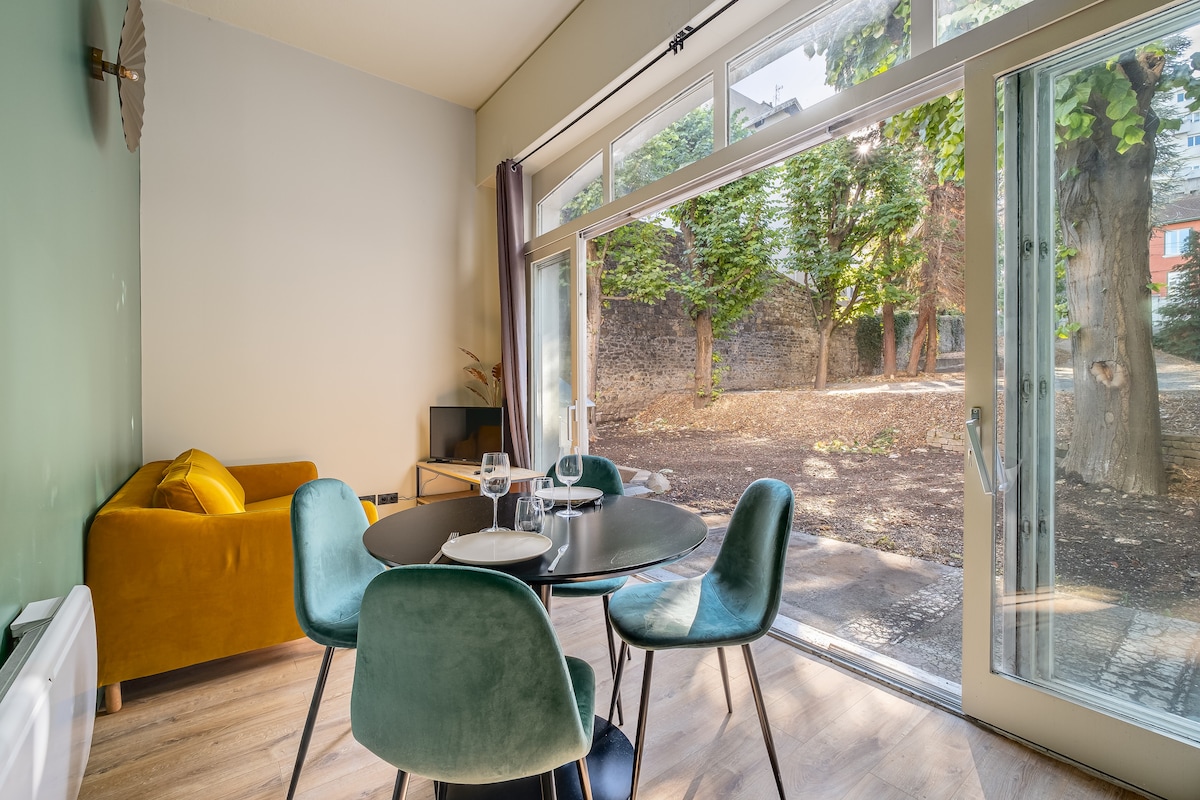 A spacious dining area features a round table surrounded by four upholstered chairs. A vibrant sofa is visible in the background, complementing the modern decor. Large glass doors open directly to a serene outdoor space with greenery, enhancing the room's natural light.