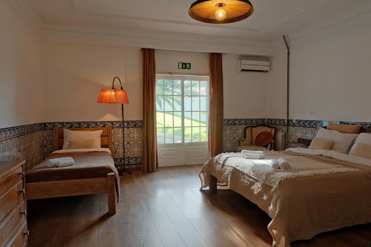 The spacious room features two beds arranged comfortably, with a warm-toned bedding set. A wooden dresser stands against the wall, and a cozy armchair sits nearby. Large windows allow natural light to brighten the space, with views of greenery visible outside.