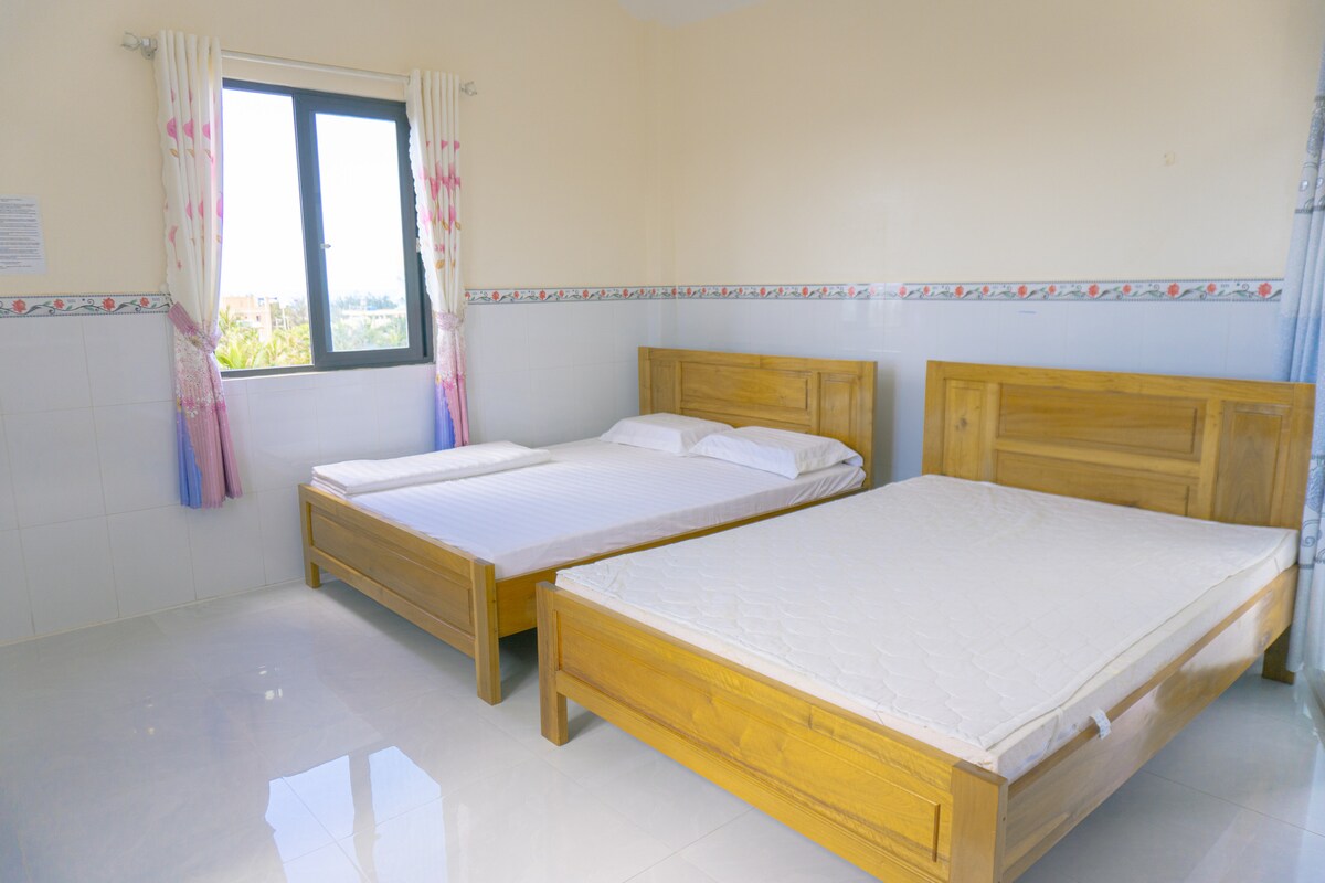 A well-lit bedroom is furnished with two wooden beds, each featuring a simple design. Light streaming in through the window highlights the clean, neutral walls. Soft pastel curtains frame the window, adding a touch of color to the space. A serene atmosphere is created by the uncluttered layout.