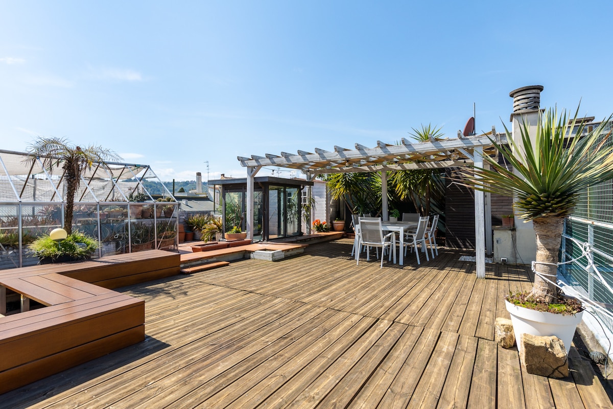 A spacious terrace is shown, featuring wooden decking and various potted plants. A pergola provides shade for outdoor seating, which includes a table and chairs. A small lounge area is present, and the skyline is visible in the background, adding character to the space.