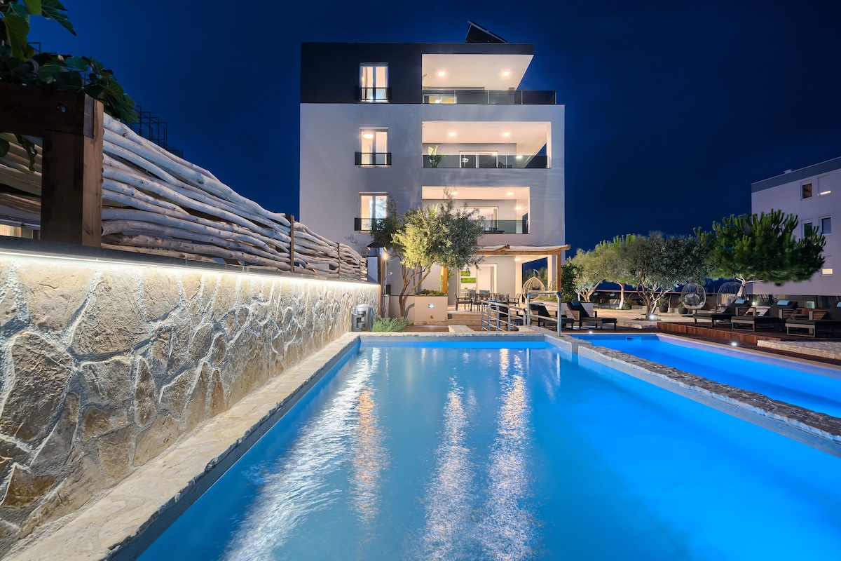 The exterior features a modern three-story building, highlighted by illuminated balconies and surrounded by lush greenery. Two inviting pools are visible in the foreground, with crystal-clear water reflecting the night sky, creating a serene retreat atmosphere.