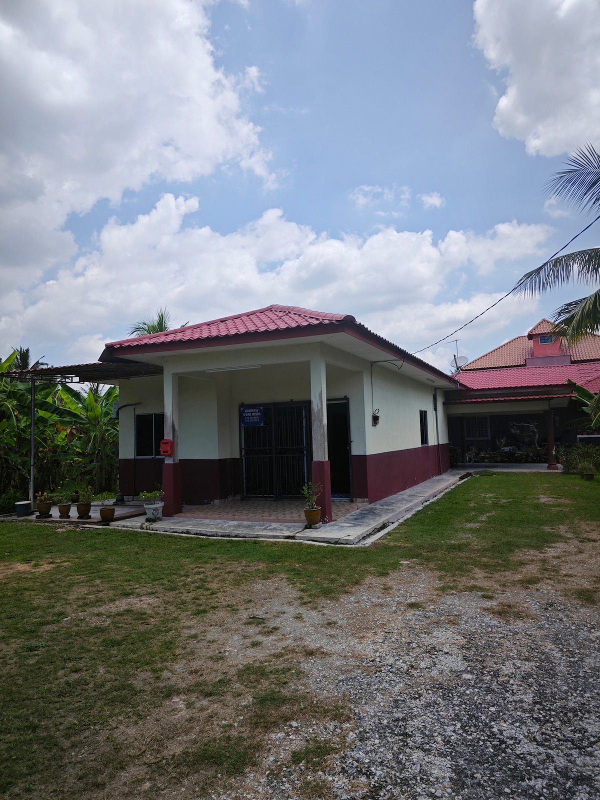 A single-story house is set in a spacious yard, surrounded by greenery. The façade features a combination of white and maroon paint, with a red-tiled roof. Potted plants line the entrance, and a walkway leads to the door, providing access to the interior.