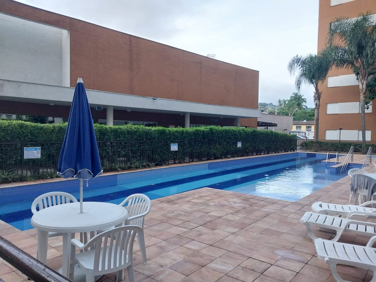 A swimming pool area is depicted, featuring a blue water surface and surrounded by a paved patio. Comfortable loungers and a round table with chairs are arranged nearby. A large umbrella provides shade, and decorative hedges line the perimeter, creating a serene environment.