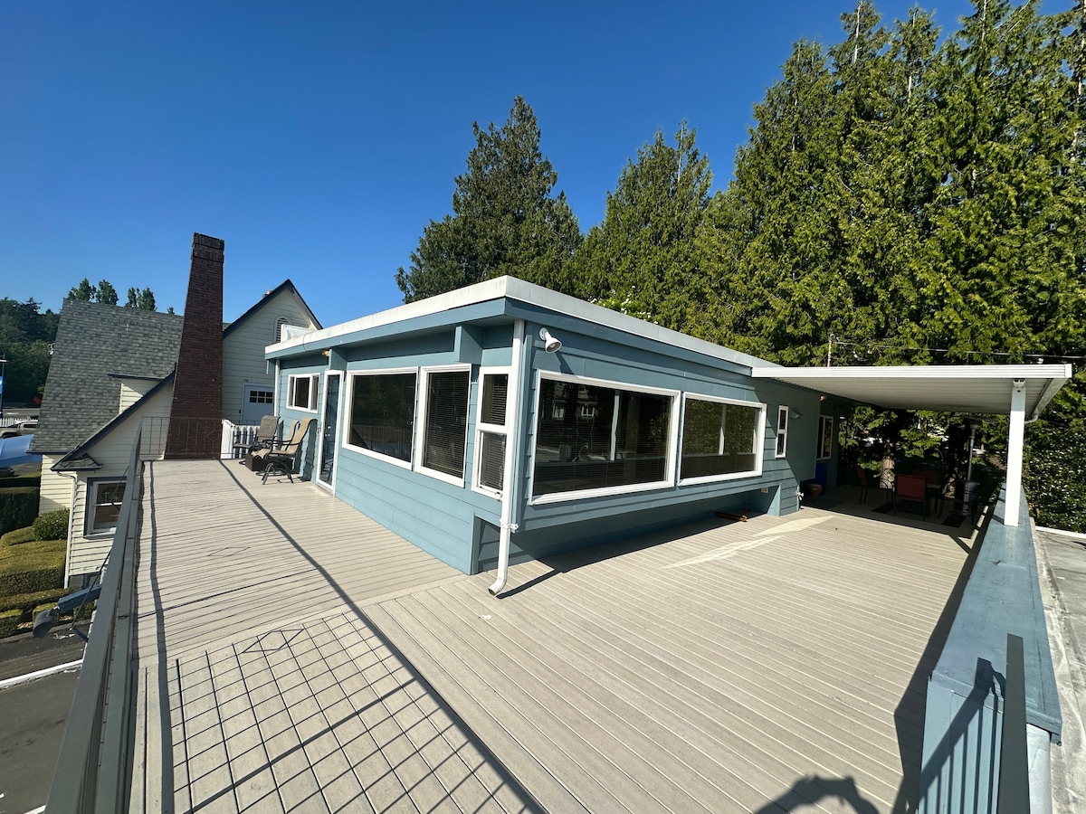 A spacious deck features a light-colored wooden surface and is surrounded by large windows that invite natural light. Several chairs are arranged comfortably, providing ample space for relaxation. Lush trees provide a backdrop, enhancing the serene setting.