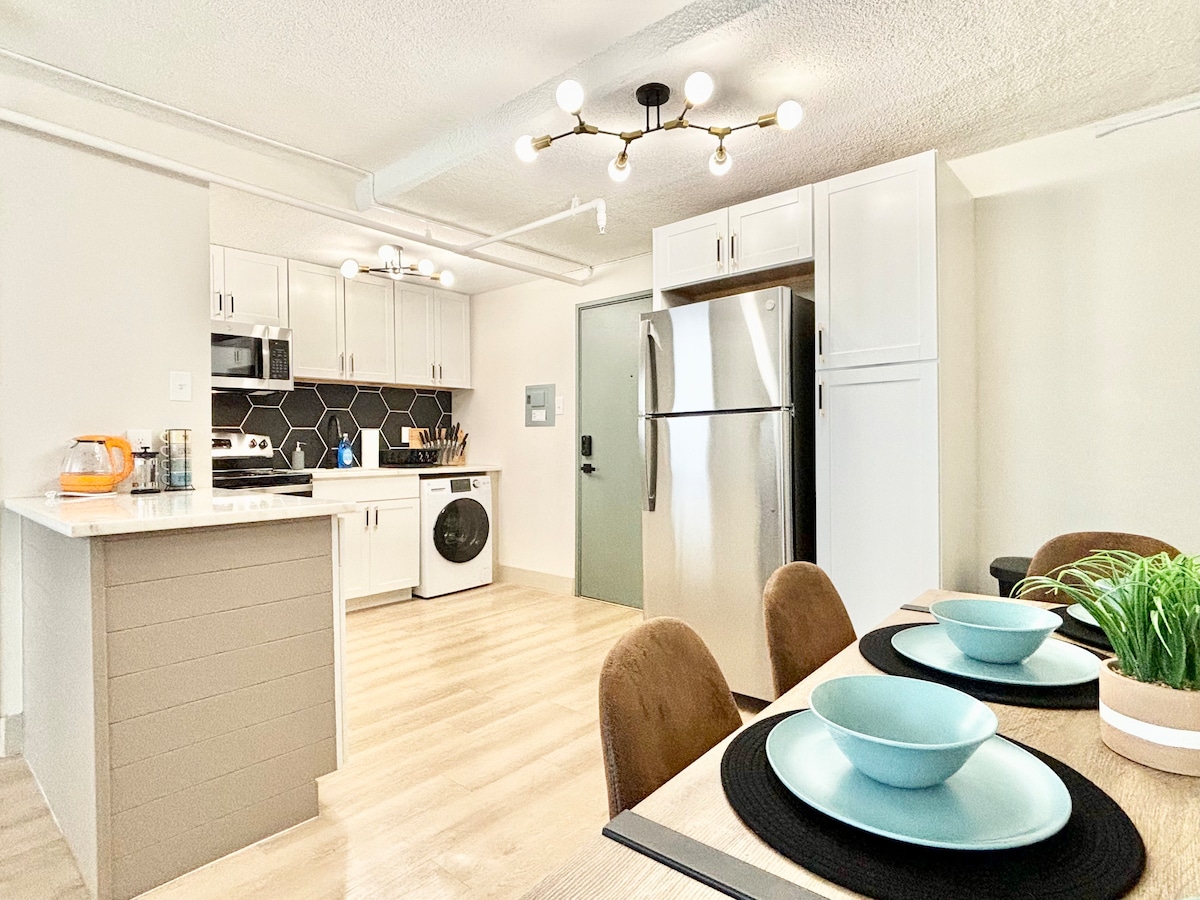 A modern kitchen is presented with sleek white cabinets and a stainless steel refrigerator. A dining area features a wooden table set with turquoise plates and a small plant centerpiece. An open layout allows for easy movement and natural light to fill the space.