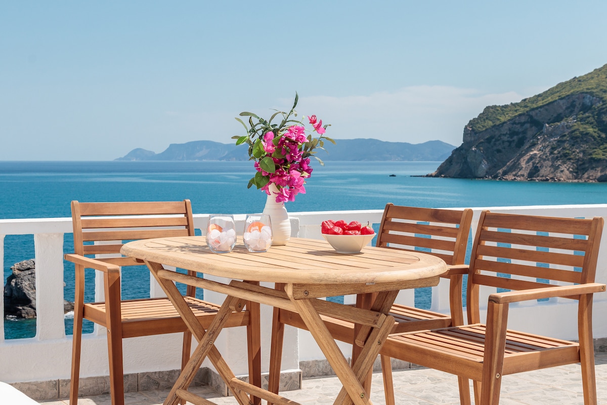 A wooden dining table is set on a spacious patio, surrounded by three matching chairs. A vase filled with pink flowers adds a vibrant touch, while a bowl of red fruit and two glasses are positioned on the table. The peaceful sea and distant hills create a scenic backdrop.