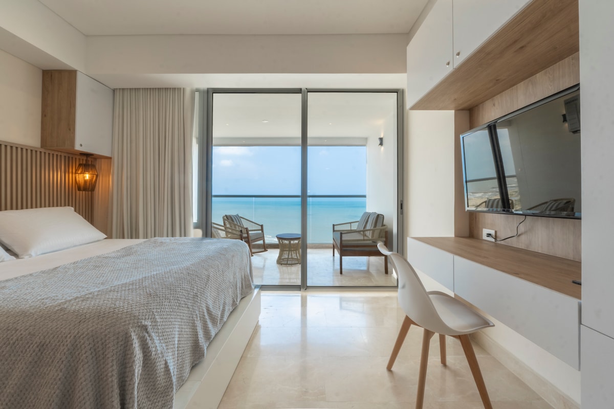 A bright bedroom is designed with modern furnishings, featuring a spacious bed dressed in neutral linens. A large glass sliding door offers unobstructed views of the ocean, leading to a furnished balcony. A flat-screen television is mounted on a wooden cabinet, providing ample storage.
