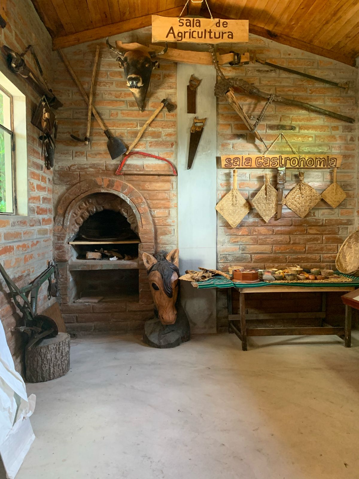 An agricultural room is presented with a brick wall adorned with various traditional farming tools. A stone oven is visible, alongside a wooden horse head ornament. On the table, items related to indigenous cuisine and craftsmanship are displayed, enhancing the cultural ambiance of the space.