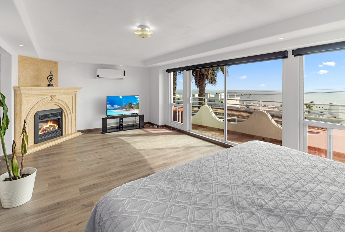 A spacious primary bedroom features a king size bed with a premium comforter. A large glass door opens to a balcony with ocean views. A modern fireplace and a massive Smart TV are included, enhancing the room's functionality and comfort.