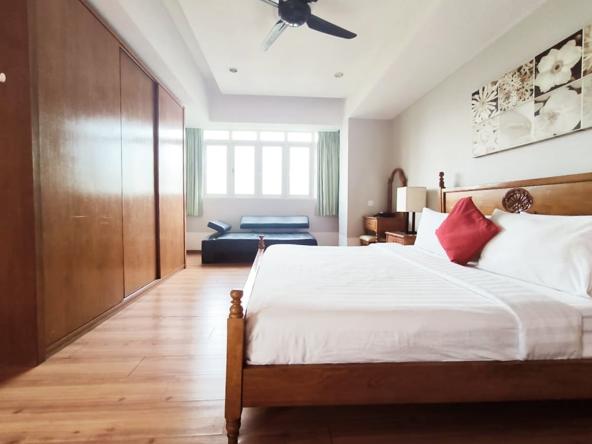 A spacious bedroom features a wooden bed with a white bedding and a decorative red pillow. Light wooden flooring complements the room. A bright window allows natural light to fill the space, and a dark sofa is positioned in the corner. Built-in wardrobes form one wall.