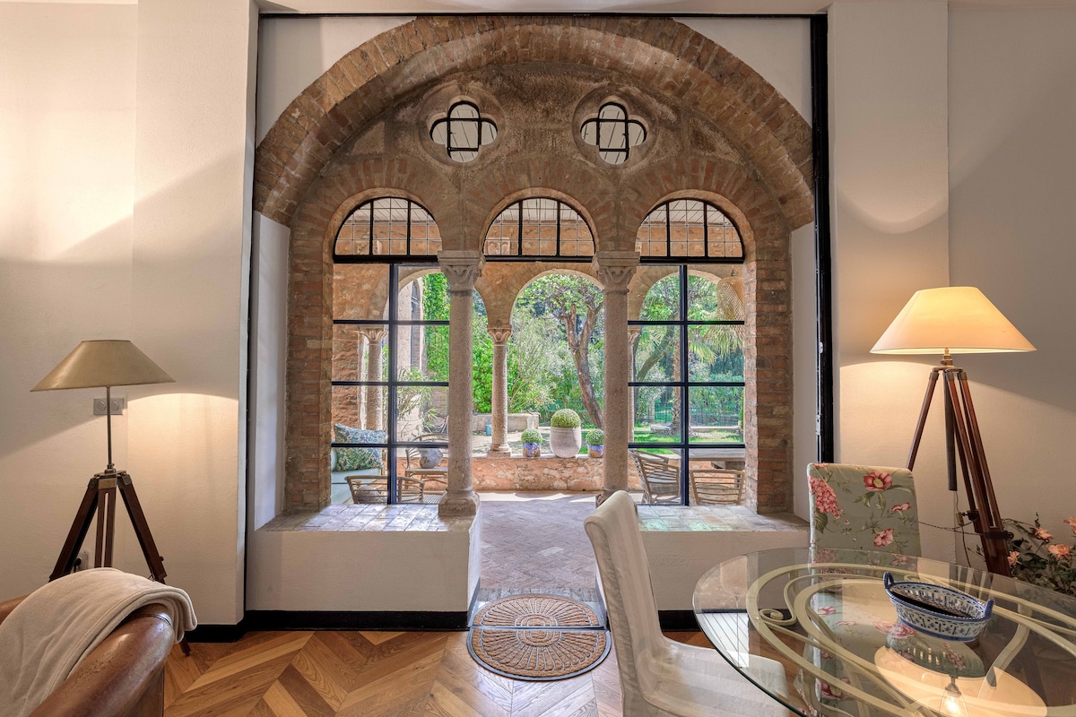 An interior view captures an elegant archway framed by tall windows, leading to a serene garden. Light filters through the panes, highlighting the finely crafted brickwork. A stylish dining area with a round glass table and flowering decorative elements is featured, along with soft lighting from nearby lamps.