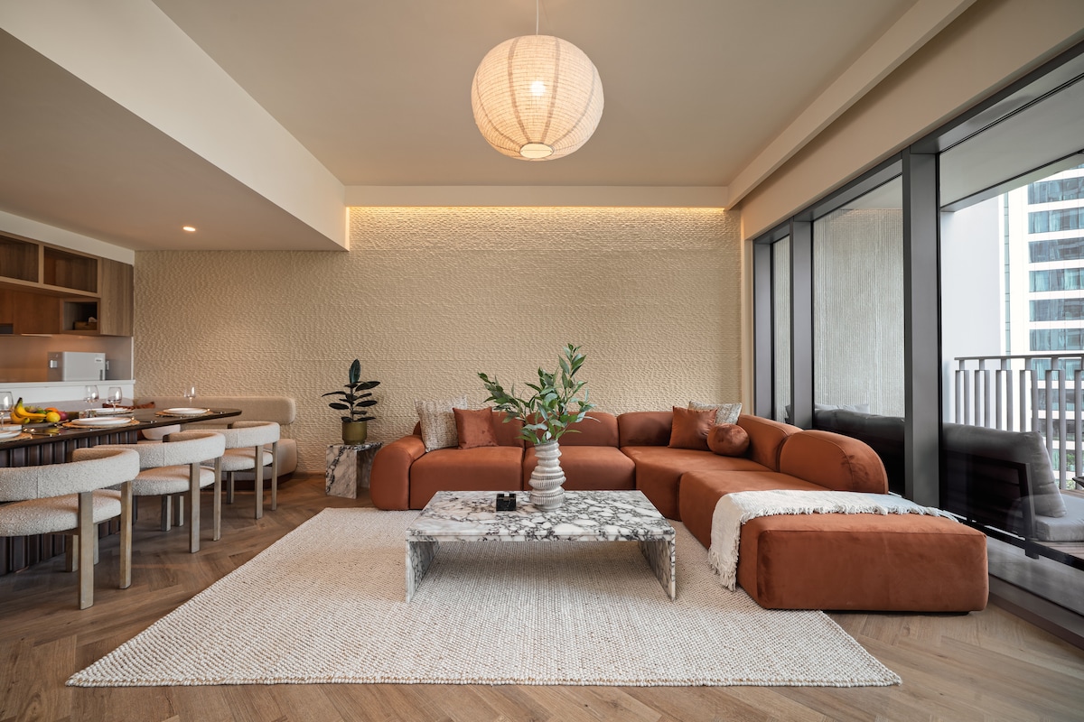 The spacious living area features a custom-made sofa in warm tones, complemented by a stylish marble coffee table. A dining area with seating for eight is visible, along with contemporary light fixtures, natural hues, and large windows offering views of the outdoor balcony.