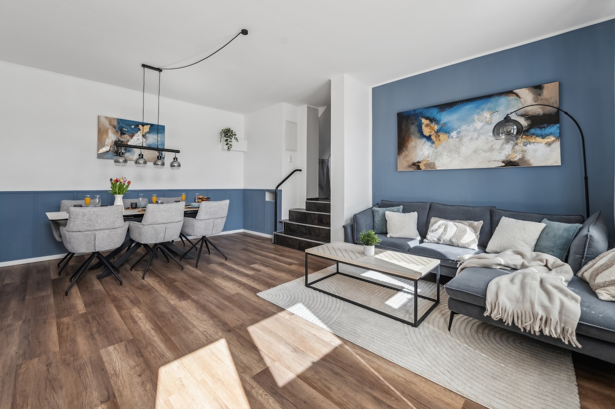 A spacious living and dining area features a modern dining table set for six, illuminated by pendant lights. A comfortable seating arrangement with soft cushions is positioned adjacent to a coffee table, while artwork adorns the blue accent wall. Natural light fills the space through large windows.