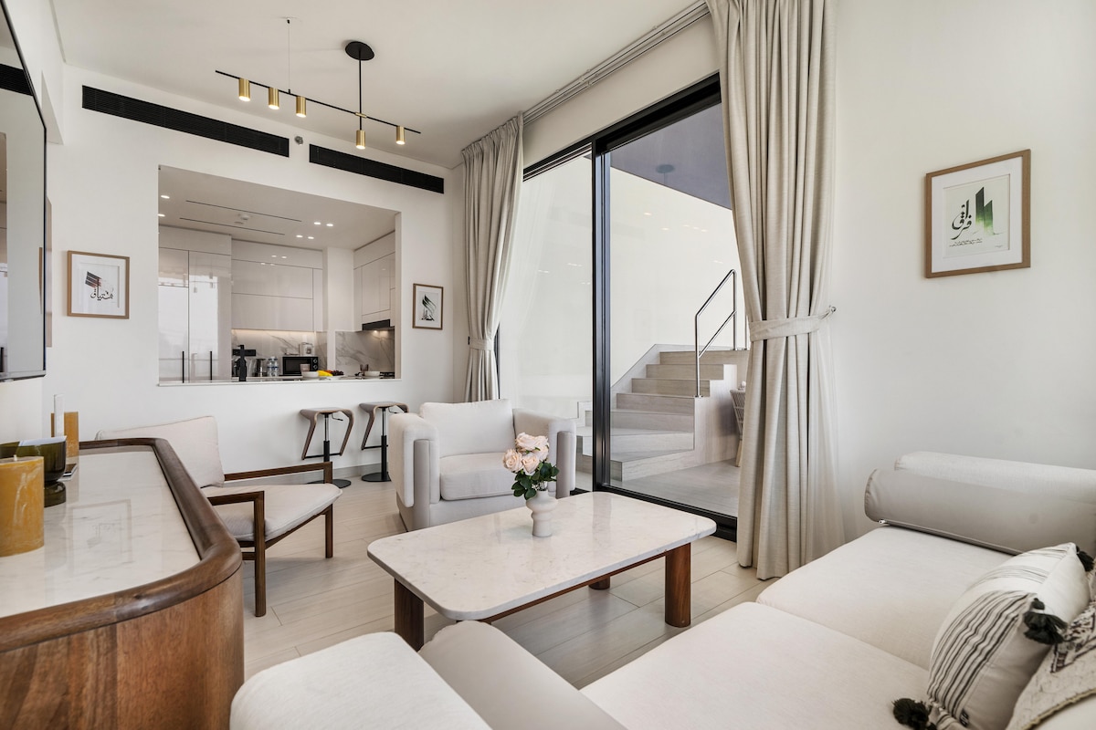 A well-appointed living area features a modern sofa and a sleek coffee table. Natural light pours in through large glass doors, leading to an outdoor space. Soft neutral tones are present, complemented by tasteful decor elements, creating a serene environment for relaxation.