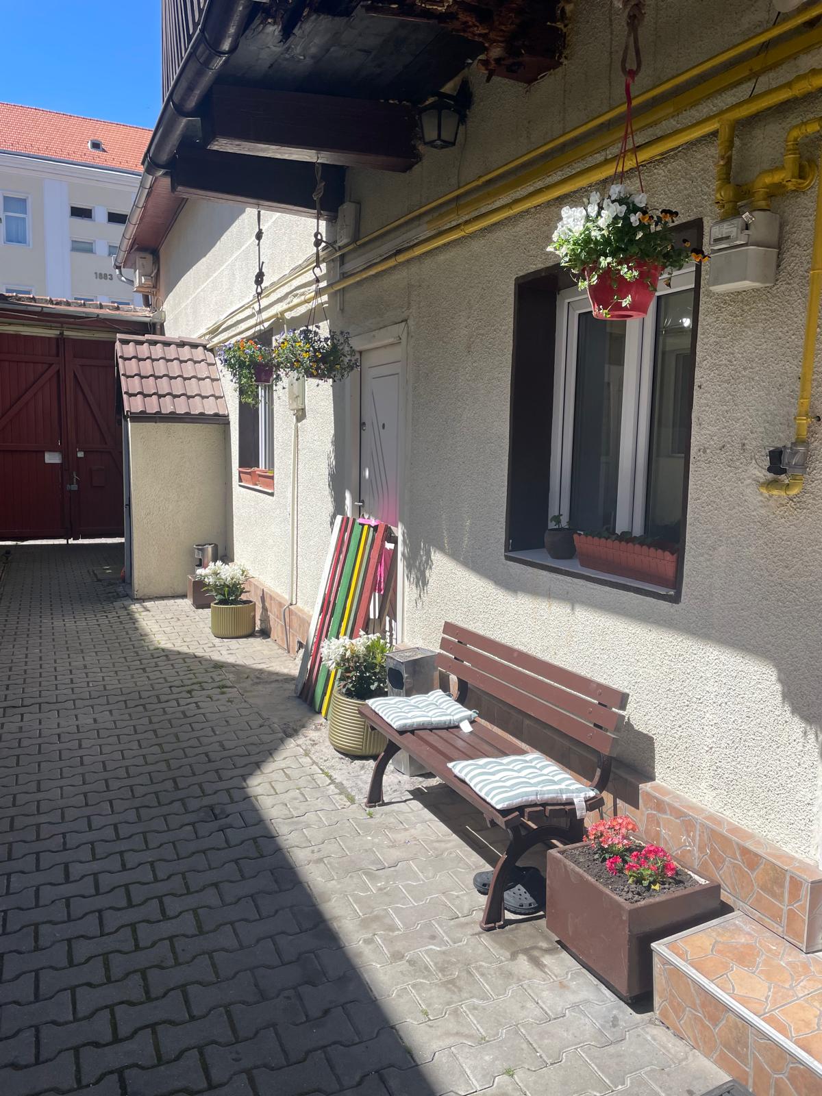 A shaded outdoor area features a wooden bench adorned with cushions. Colorful planters filled with flowers are arranged along the wall, while hanging pots add greenery above. The stone-paved pathway leads to a red door, providing an inviting atmosphere for relaxation.