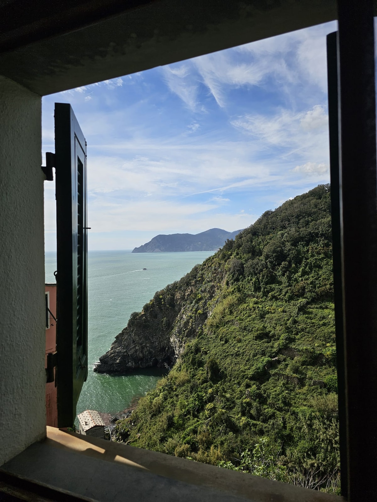 A view is captured through an open window, showcasing a serene coastal landscape with lush green hills descending into the blue sea. The horizon is marked by distant mountains under a partly cloudy sky, enhancing the tranquil outdoor scenery.