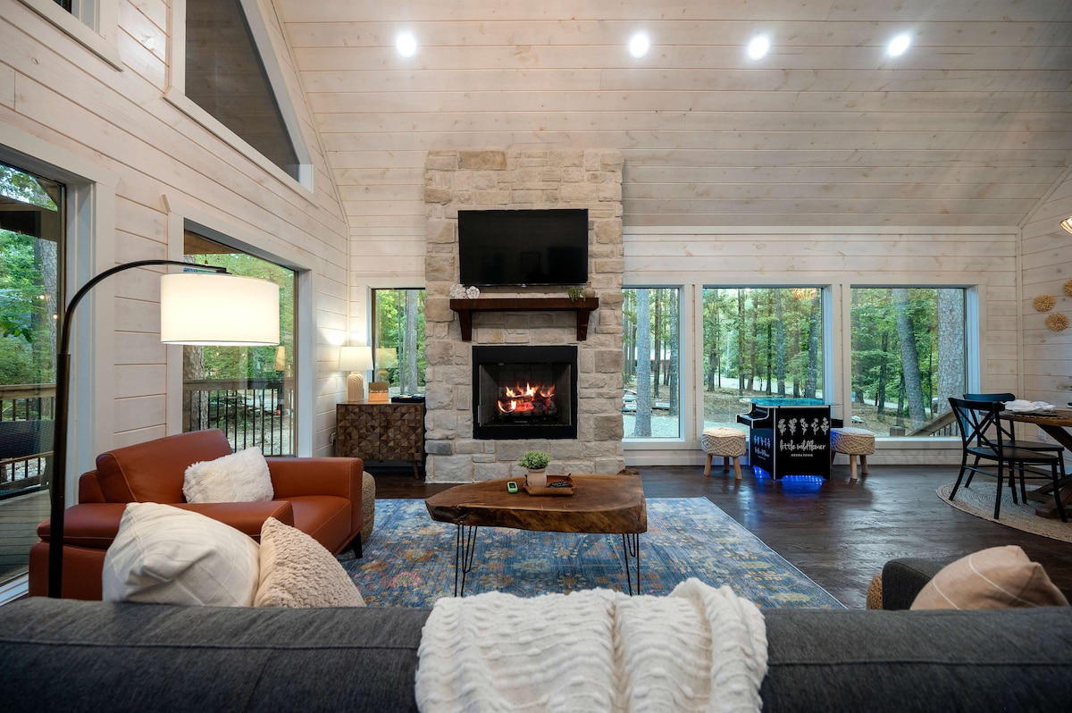 A cozy living space features a stone fireplace with a mounted TV above it. Large windows frame the room, providing views of the surrounding woods. Comfortable seating includes a leather chair and a plush sofa, accompanied by a unique coffee table on a patterned rug.