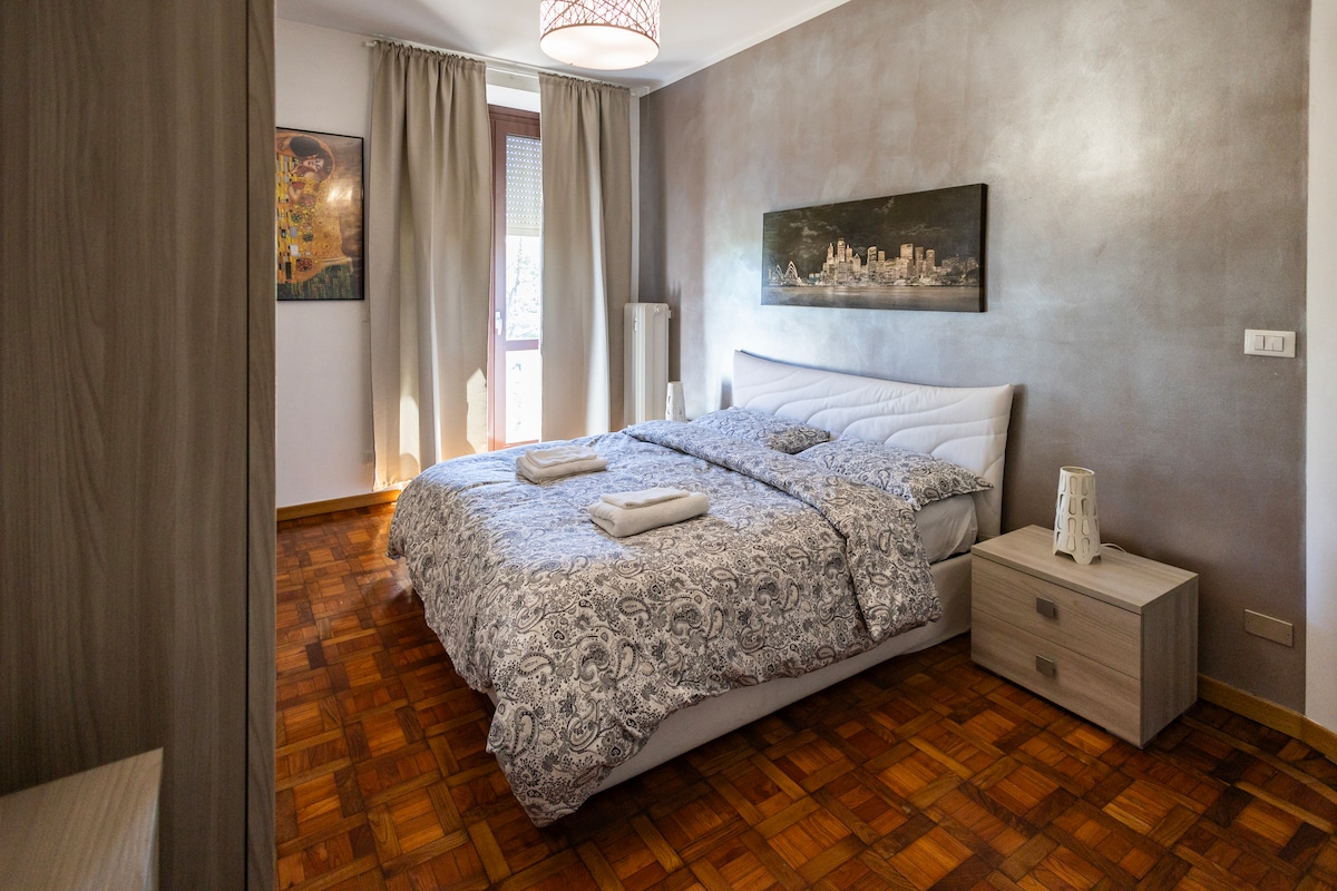 A spacious bedroom features a large bed with a patterned duvet, complemented by neat linens. Natural light enters through a window adorned with sheer curtains. A bedside table and a light fixture provide functionality and style, while artwork adds a decorative touch to the walls.