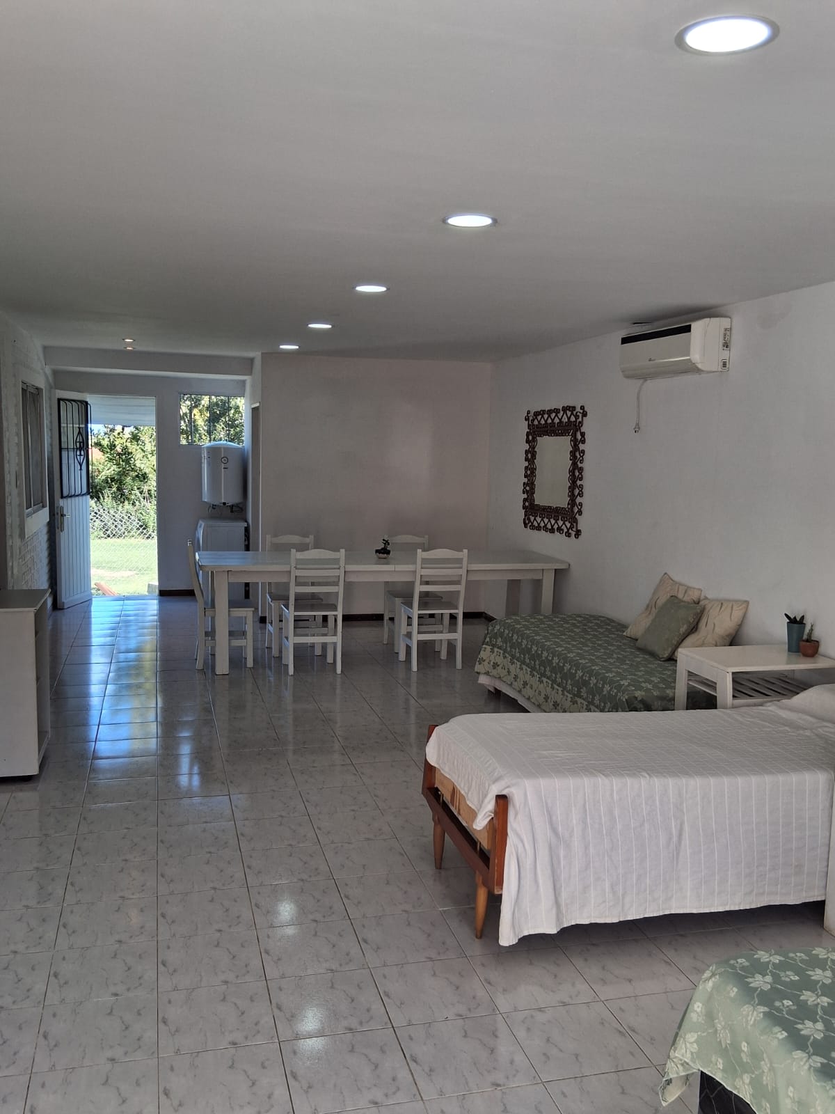 A spacious living area features a comfortable seating arrangement and a dining table set for several individuals. Natural light enters through an open doorway, illuminating the white walls and tiled floor. Air conditioning and a wall mirror enhance the practical design.