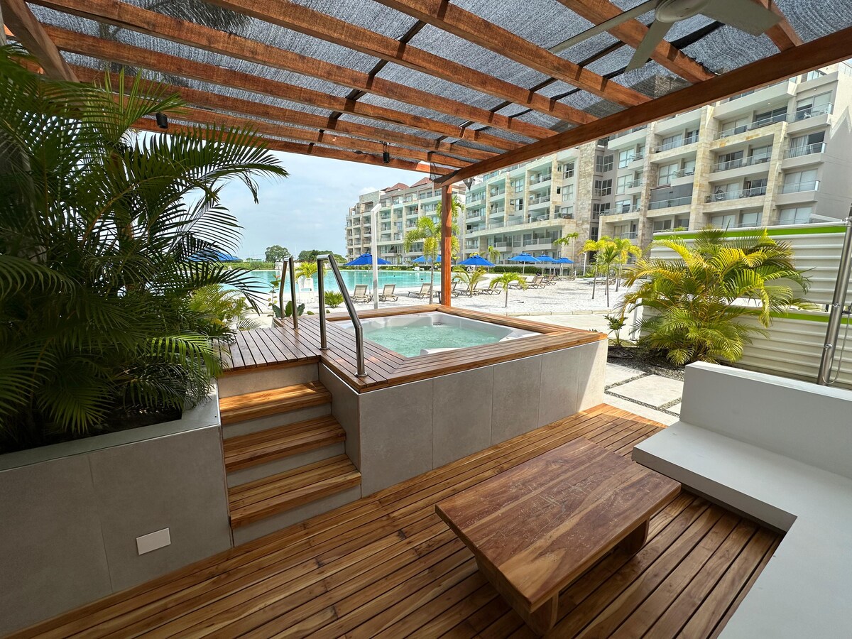 A private outdoor space features a hot tub surrounded by tropical plants and wooden decking. Steps lead from the tub to the sandy beach area visible in the background. Sun loungers are arranged on the beach, complementing the serene atmosphere.