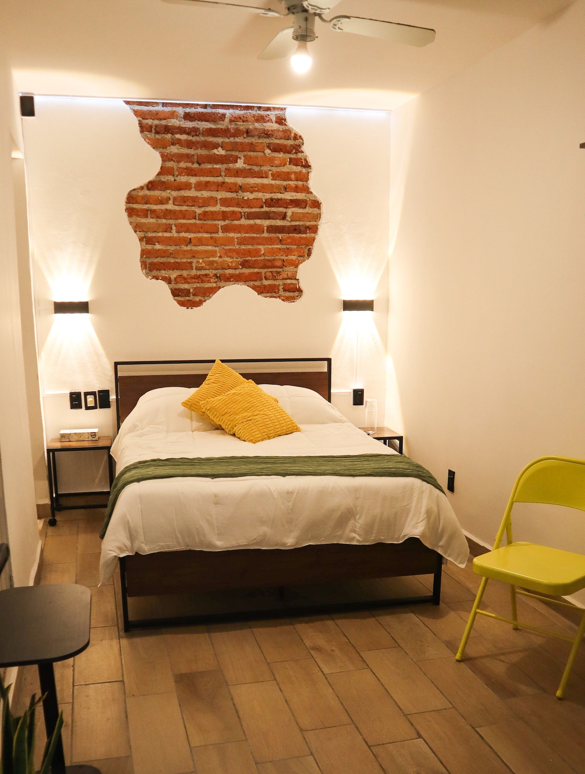 A cozy guest room features a spacious bed with white linens and a bright yellow accent pillow. Exposed brick walls provide a textured backdrop, while bedside lamps offer soft light. A small yellow chair adds a pop of color, and warm wooden flooring enhances the inviting atmosphere.