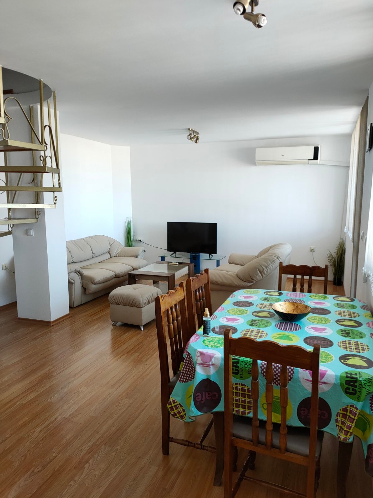 An open living area is depicted, featuring a light-colored sofa set and a small television on a stand. A dining table with a multicolored tablecloth and wooden chairs is present, enhancing the room’s functionality. Large windows allow natural light to fill the space.