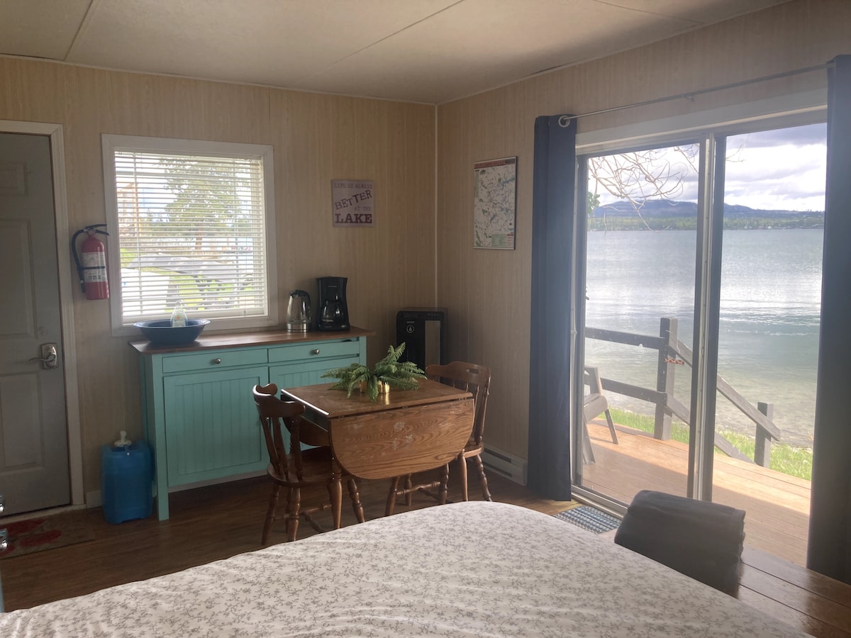 A cozy interior features a small wooden dining table with four chairs beside a blue sideboard. A coffee maker and kettle are visible. Large sliding glass doors showcase scenic lake views and access to a private deck. Natural light brightens the space.